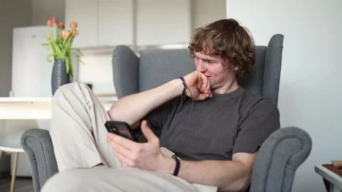 Smiling while looking at his smartphone, a young man sits comfortably in a chair Stock Footage 302633935