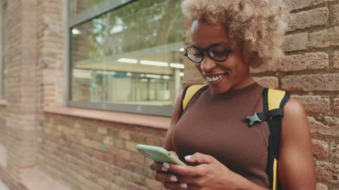 Smiling woman with backpack writes sms on mobile phone while standing outside Stock Footage 252031449