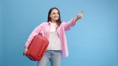 Smiling woman doing advertisement in studio on blue background with red suitcase 스톡 동영상 240936664