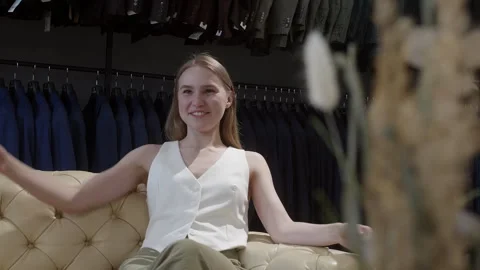 Smiling woman looking away while sitting on sofa in men's clothing store Stock Footage 303459348