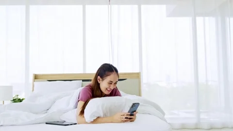 Smiling woman lying on a bed in a bright bedroom, using a smartphone and relax 動画素材 323065853