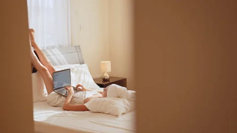 Smiling woman lying on white bed while lifting legs up and using app on Laptop Stock Footage 166118501