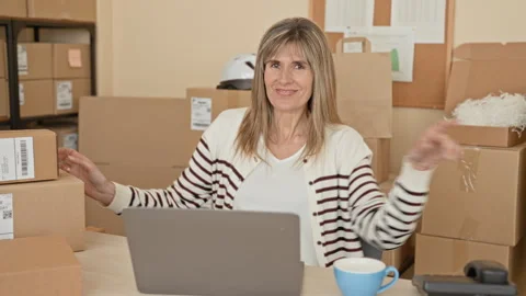 Smiling woman points finger at camera amid stacked parcels and laptop in bu.. Stockbeeldmateriaal 337432850