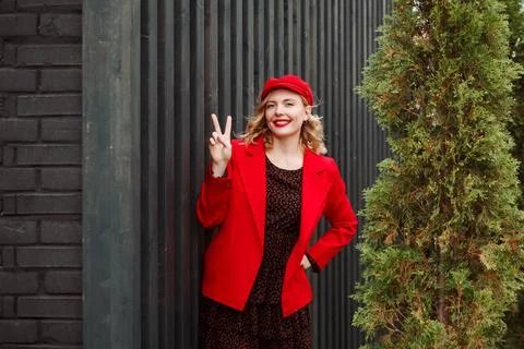 Smiling woman in red jacket and cap is showing victory symbol in front of dark Stock Photos