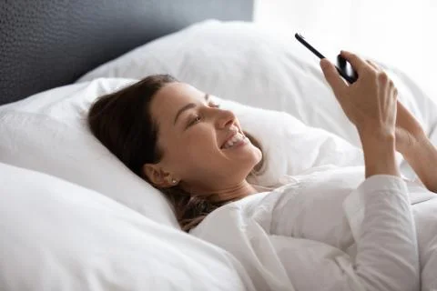 Smiling woman relaxing in bed, reading sms with good news. Stock Photos