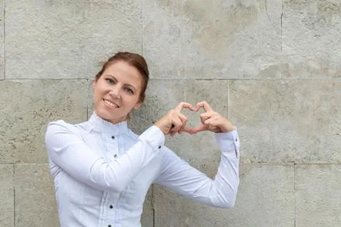 Smiling woman showing heart sign on urban wall background with copy space Stock Photos