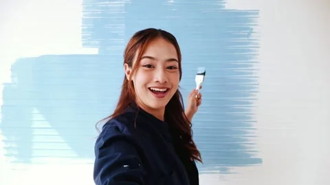 Smiling woman takes vlog while painting wall light blue with a brush. Stock Footage 311566176