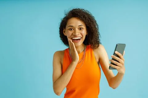 Smiling woman touching cheek while holding mobile phone over blue studio Stock Photos