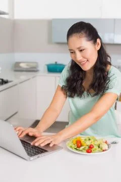 Smiling woman using laptop while having salad in kitchen Stock Photos