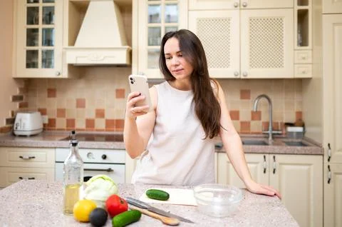 Smiling woman using smartphone while preparing fresh vegetable salad in cozy  Stock Photos