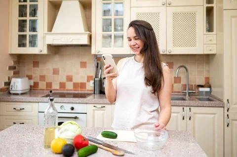 Smiling woman using smartphone while preparing fresh vegetable salad in cozy  Stock Photos