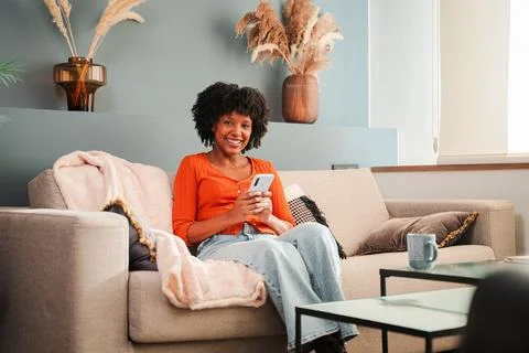 Smiling woman using smartphone while sitting comfortably on a soft couch in a Stock Photos