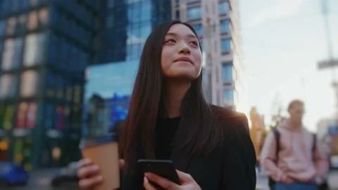 Smiling woman walking while using phone hold coffee in urban city buildings look Stock Footage 156737184