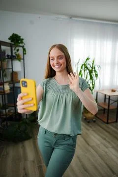 Smiling woman waves while taking a selfie in bright living room Stock Photos