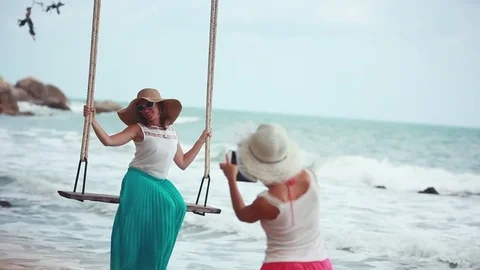 Smiling woman wearing hat taking photo with phone her friend on a swing at beach Stock Footage