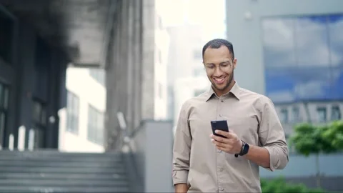 A smiling young adult man is using a smartphone while walking on the street near Stock Footage 234068816