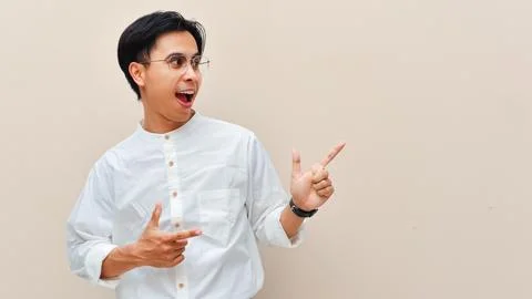 Smiling young Asian man pointing finger to the side at copy space against a.. Stock Photos