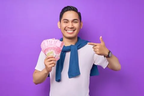 Smiling young Asian man wearing white t-shirt pointing fingers at cash mone.. Stock Photos