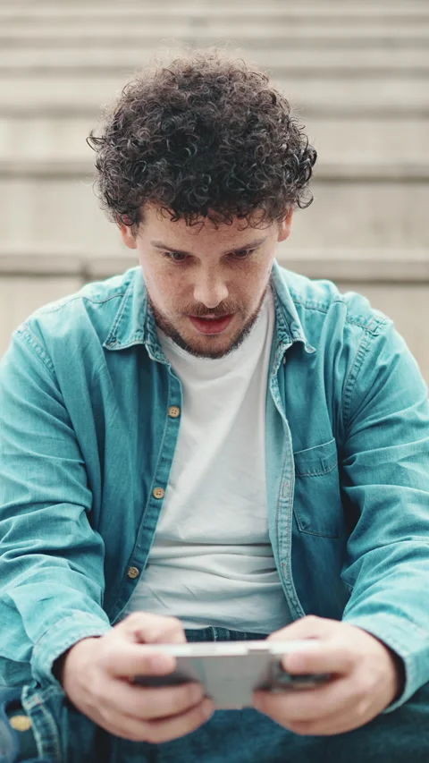 Smiling young bearded man in denim shirt sitting on high steps and using mobile Stock Footage 227005186