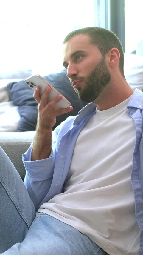 Smiling young bearded man sitting on the floor at home, happily sending a voice Stock Footage 345115147