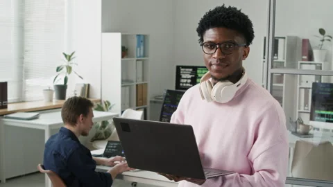 Smiling Young Black IT Developer with Headphones Holding Laptop in Office Video stock 330126854