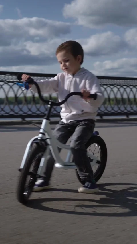 Smiling young boy rides a white balance bike along a sunny waterfront path under Stock Footage 347031395
