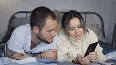 Smiling young couple in the bed late and taping on their smartphones Stock Footage 128546384