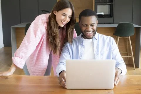 Smiling young couple using laptop together at home kitchen table while work.. Stock Photos