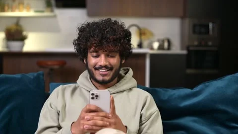 Smiling young curly Indian guy using smartphone sitting on the couch at home Video stock 176394302