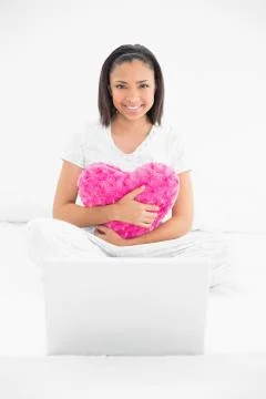 Smiling young dark haired model cuddling a pillow and using a laptop 스톡 사진
