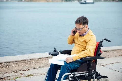 Smiling young disabled man use phone on a wheelchair in a park at sunlight Stock Photos