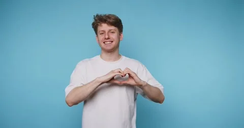 Smiling young man forms a heart shape with his hands against a bright blue .. Stock Footage 313281005
