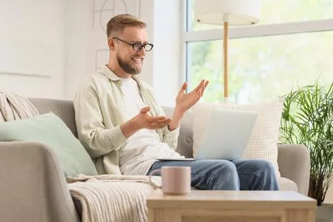 Smiling young man in glasses on sofa using laptop for video call, remote work Stock Photos