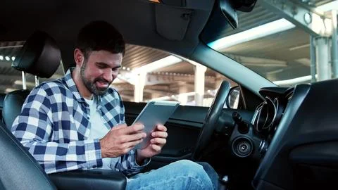 Smiling young man laughing while looking at tablet screen in the car. Transport Stock Photos