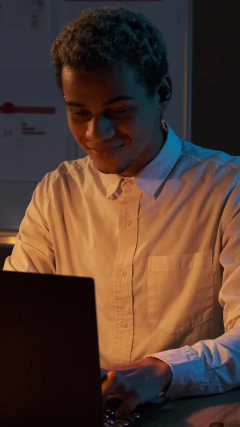 Smiling young man puts on wireless earbud while working on laptop at night. Stock Footage 326118864