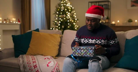 A smiling young man in Santa hat sits on sofa in the living room and unpacks Stockbeeldmateriaal 211291898