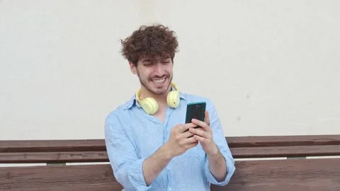 Smiling young man sitting on a bench chatting with a smartphone in a city Stock Footage 219285832