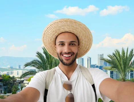 Smiling young man in straw hat taking selfie against city with buildings an.. Fotos de archivo