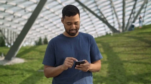 Smiling young man texting and web surfing standing on green lawn 스톡 동영상 96890264