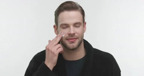 Smiling Young Man using Delicately Moisturizing Creme on Cheek, wearing Bathrobe Stock Footage 151946764