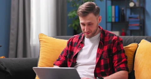 Smiling young man using laptop, sitting on couch at home, handsome male shopping Stock Footage 154044728