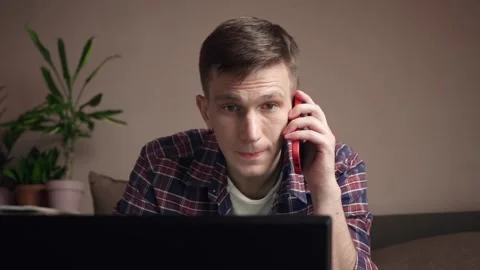 Smiling young man using laptop, talking on mobile phone while sitting on sofa. Stock Footage 211812186