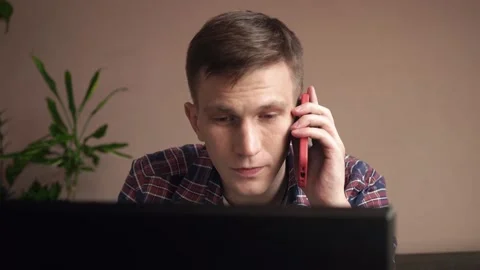 Smiling young man using laptop, talking on mobile phone while sitting on sofa. Stock Footage 212608457