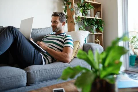 Smiling young man using a laptop on his sofa Stock Photos
