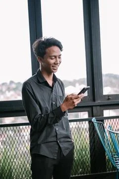 Smiling young man using smartphone near window with urban backdrop Stock Photos