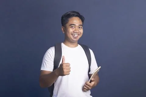 Smiling young man wearing a backpack and holding a notebook while giving a .. Stock Photos