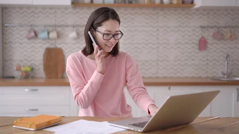 Smiling young mixed race asian business woman talking on phone using laptop sit Stock Footage 151029680