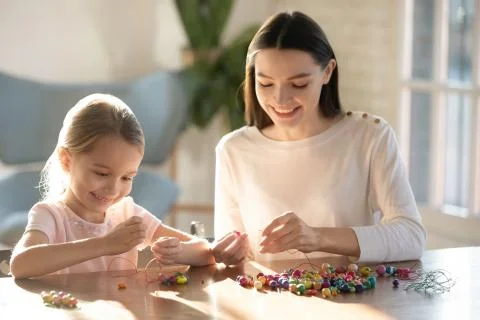 Smiling young mom stringing beads on needles with little daughter. Stock Photos