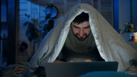 Smiling young pleasant-looking man sitting on bed covered with blanket. Guy Stock Footage 105655428