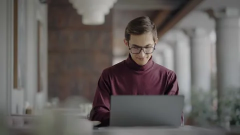 Smiling young stylish man in turtleneck and glasses working at laptop computer 库存影片 344800248